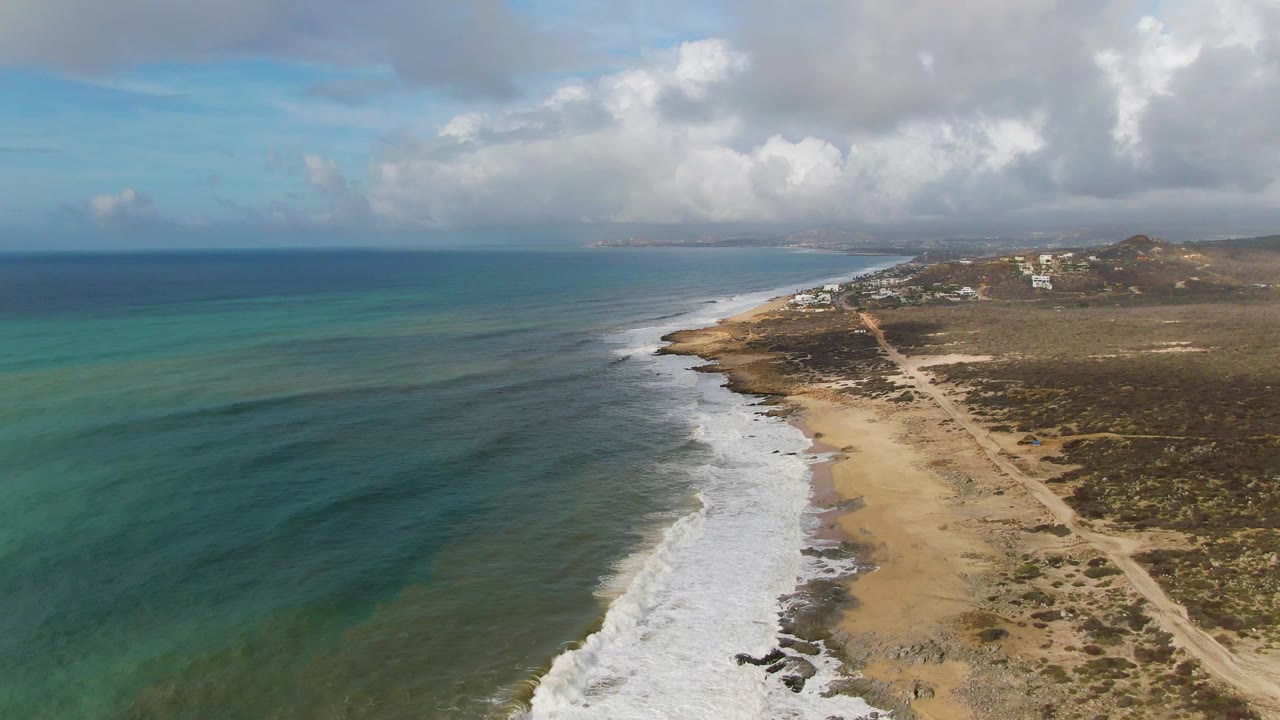 Aerial view of Punta Gorda East Cape coastline with turquoise water on the left, white surf along the sandy shore, scrubland and a dirt path on the right, and no visible people.