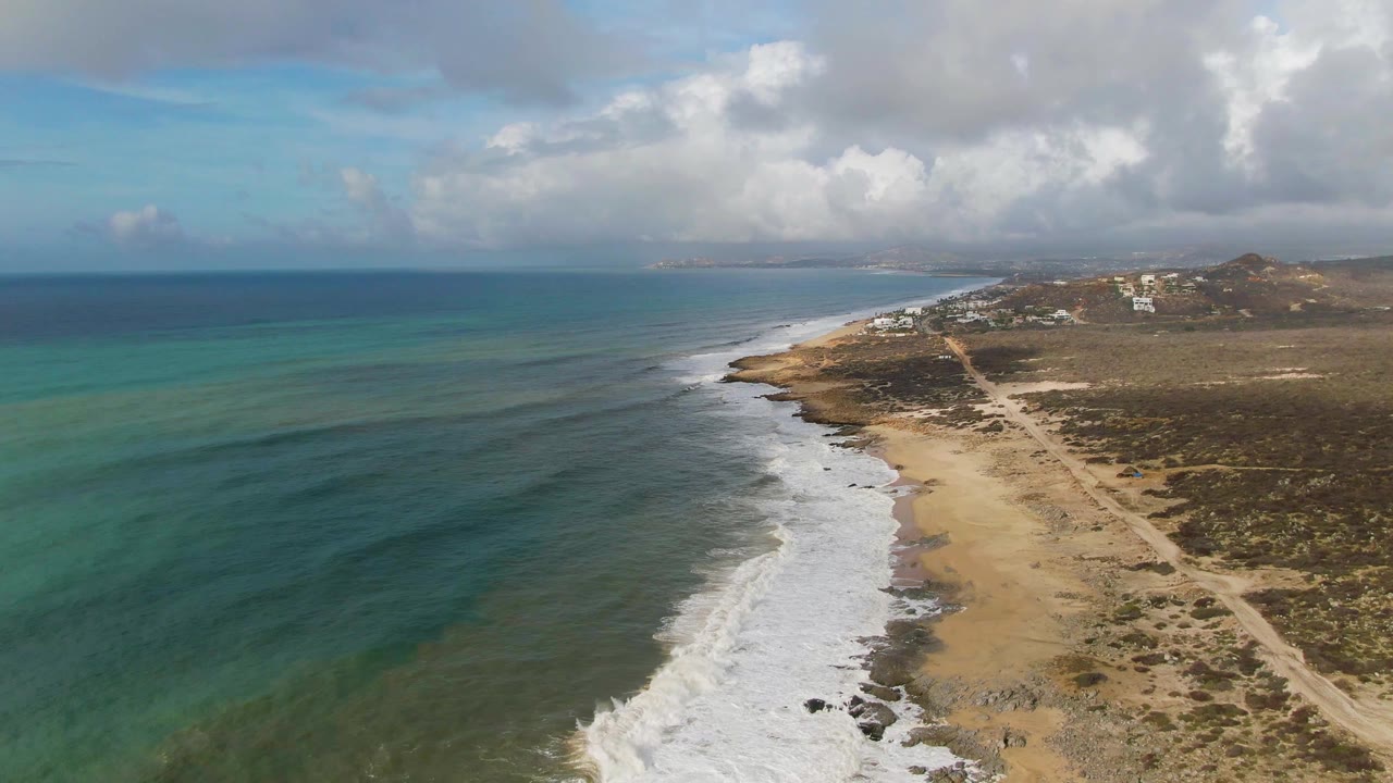 Drone view of waves breaking beside a sandy shoreline and dirt coastal road.