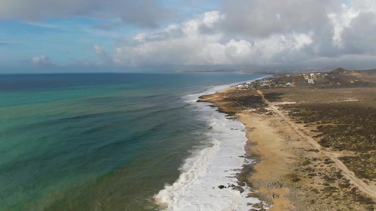 Aerial frame from Punta Gorda East Cape showing surf washing onto the sand, blue-green water extending to the horizon, dry coastal vegetation inland, distant hills, and no visible people.