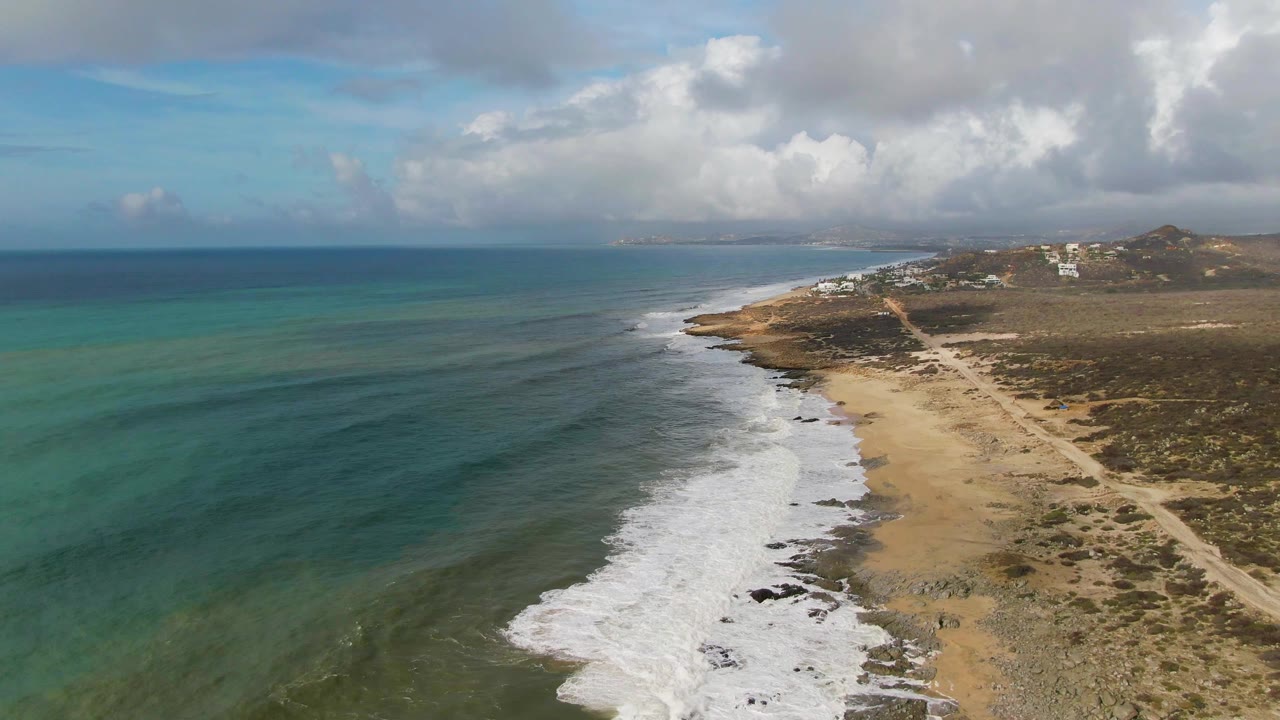 At 0:00, an aerial view looks along the Punta Gorda East Cape shoreline with turquoise water, white surf, sandy beach, sparse scrubland, distant structures, and no visible people.