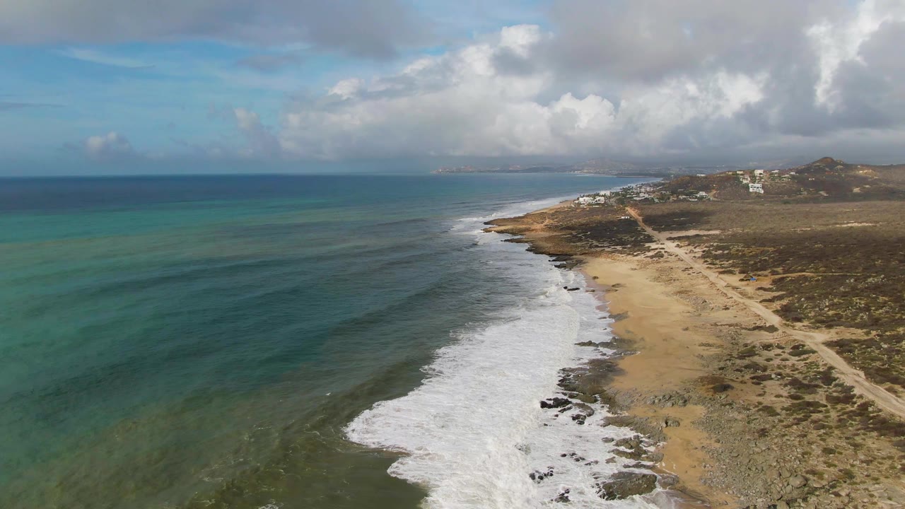 At 0:02, white waves spread across the sandy and rocky Punta Gorda East Cape shoreline while the beach path and sparse inland vegetation remain visible, with no visible people.