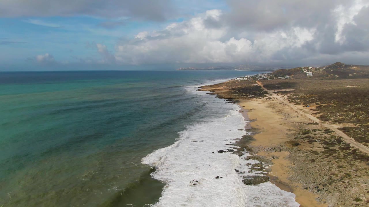 Aerial view over Punta Gorda, East Cape, Baja California Sur, with turquoise water at left, white surf on a sandy and rocky beach at right, distant hills, clouds, and no visible people.