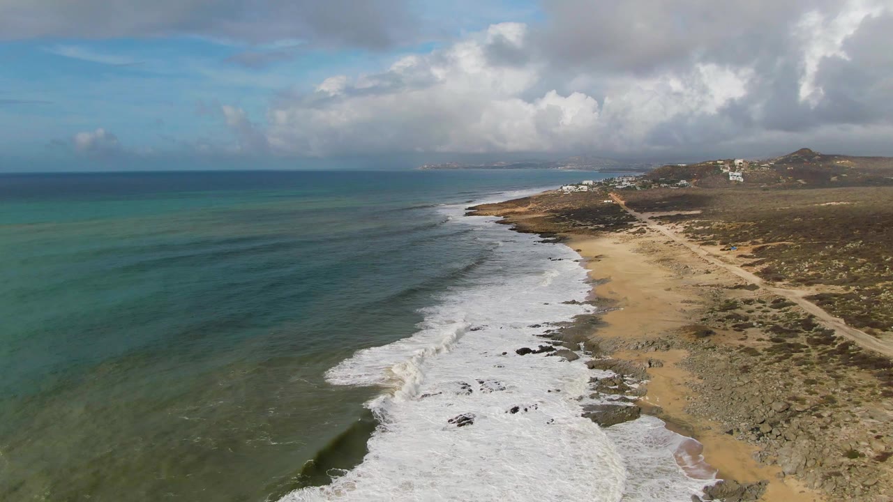At 0:02, the aerial view of Punta Gorda shows a long diagonal surf line, darker submerged patches in the water, sandy beach, scrub-covered land, clouds, and no visible people.