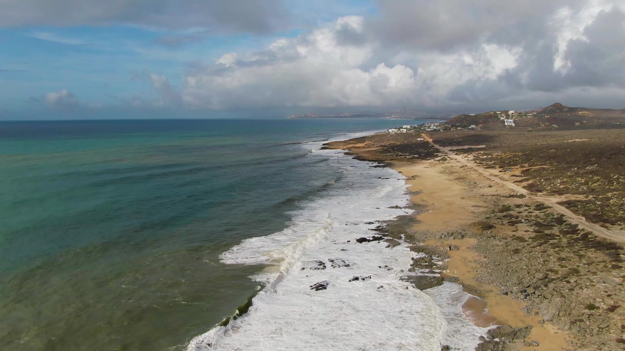 Drone view of waves washing over a broad sandy and rocky beach.