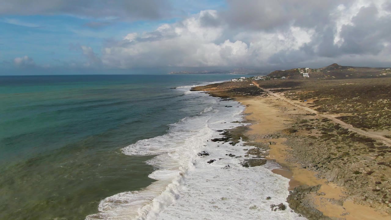 Aerial frame from Punta Gorda East Cape, Baja California Sur, showing white surf running diagonally along rocky sand, green-blue water on the left, low hills on the right, cloud cover above, and no visible people.