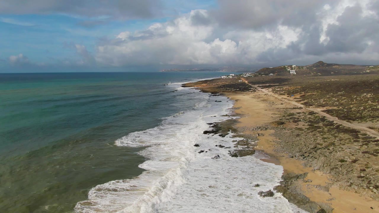 Aerial view over Punta Gorda East Cape with a wider band of white foam pushing toward the beach, turquoise shallows left of the break, dark rocks in the wash, scrubby hills inland, and no visible people.