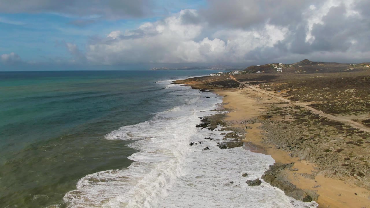 Punta Gorda East Cape coastline seen from above, with surf curling around exposed rocks near the center, tan beach and dirt tracks to the right, blue-green ocean to the left, heavy clouds overhead, and no visible people.
