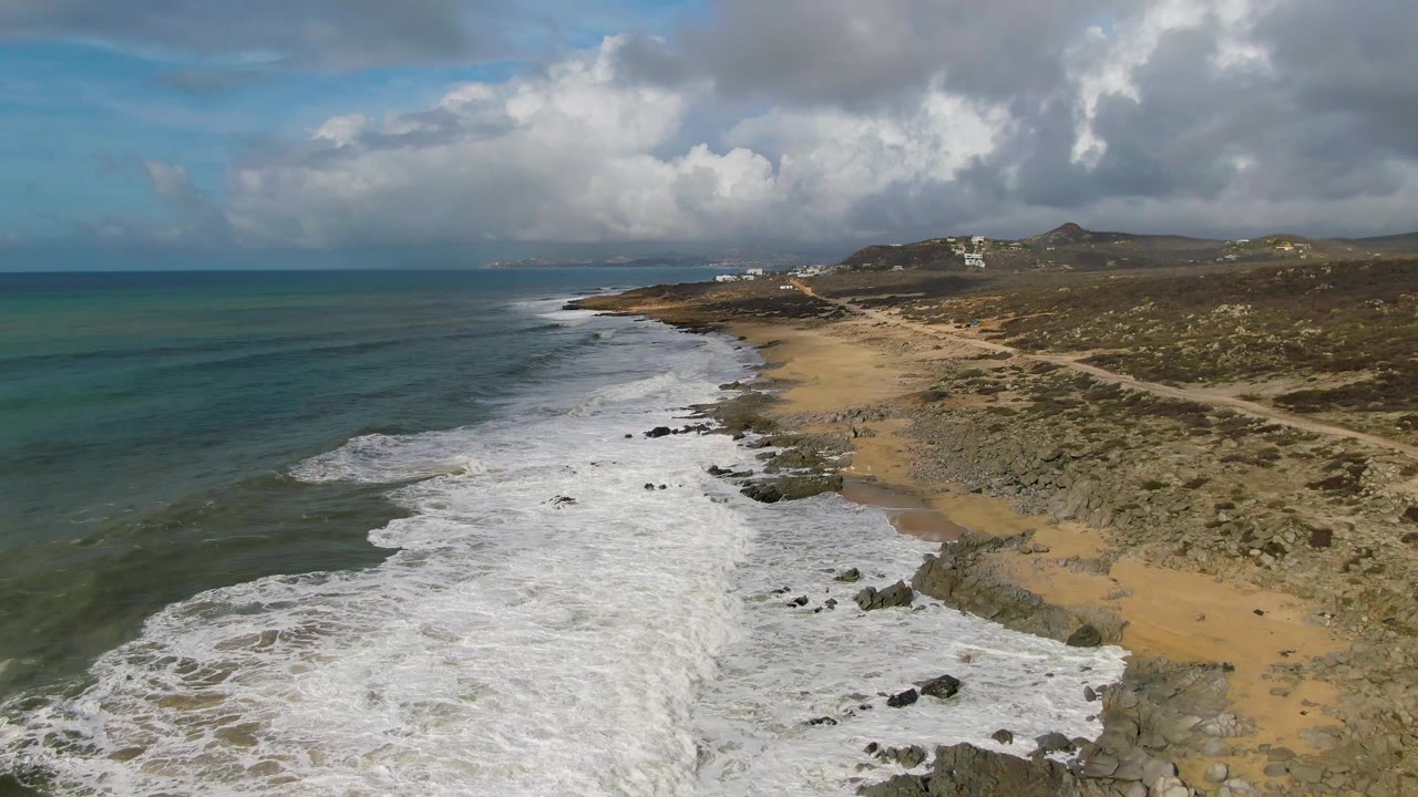 Aerial view at Punta Gorda, East Cape, Baja California Sur, with white surf reaching rocks and sand, a dirt road on the right, cloudy sky above, and no visible people.