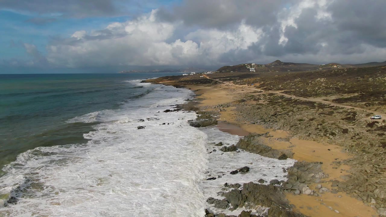 At 0:02, white water fills much of the Punta Gorda shoreline, with tan sand and boulders at center, scrub terrain and a small vehicle near the coastal road, and no visible people.