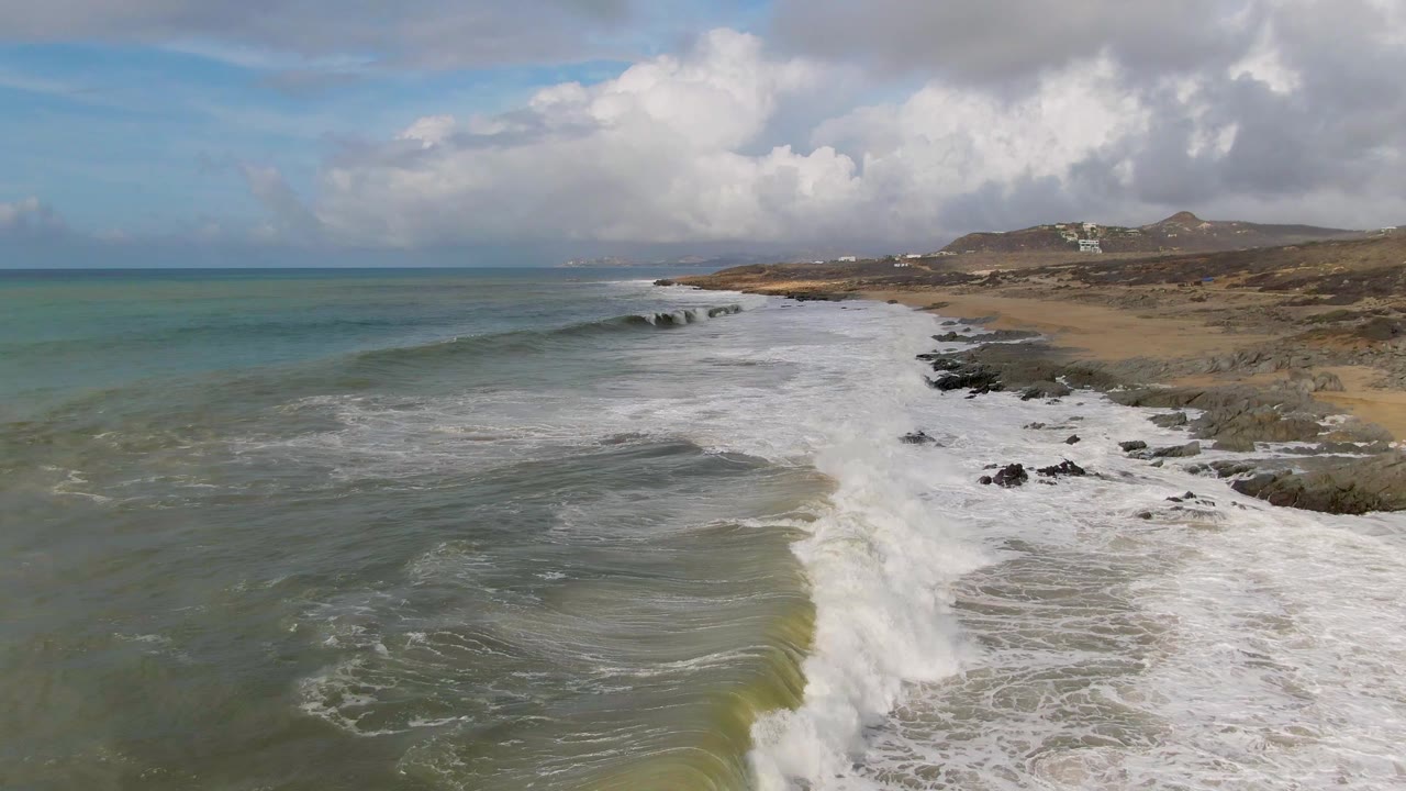 Aerial view at Punta Gorda, Baja California Sur, with green-blue ocean on the left, white surf crossing dark rocks and tan sand, low hills in back, and no visible people.