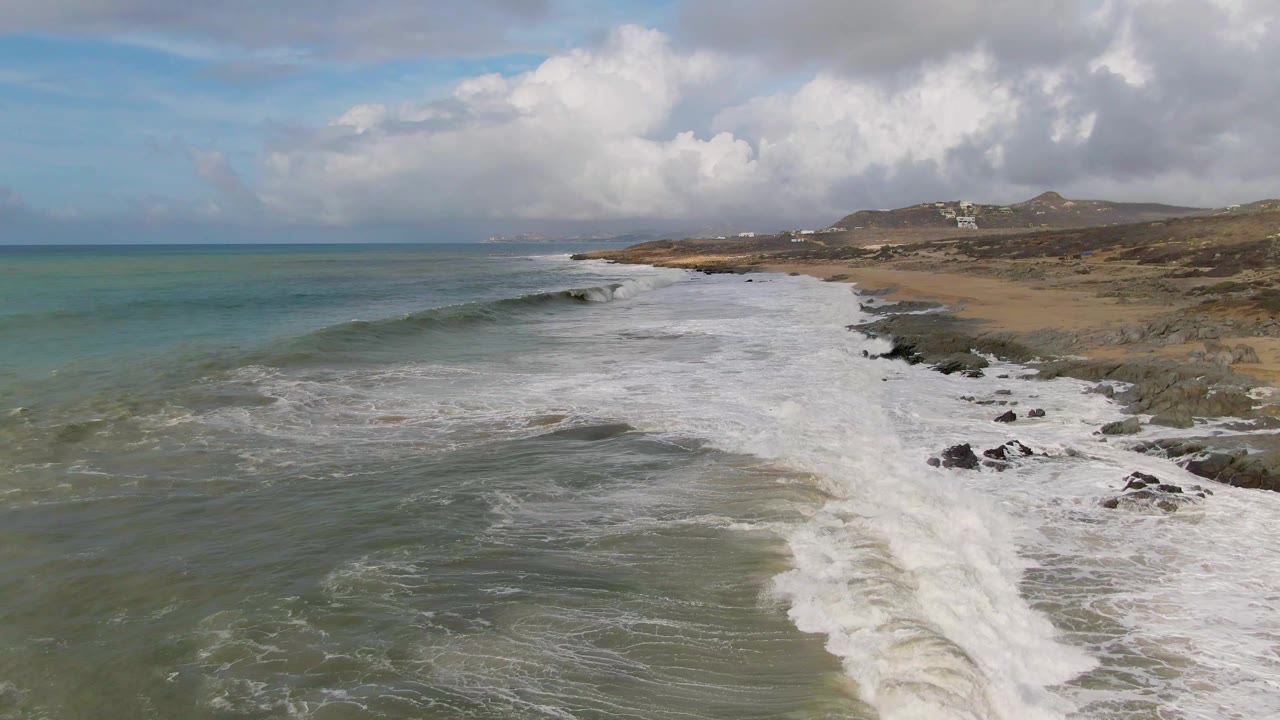 At 0:01, Punta Gorda surf forms a long white line beside the rocky shore, with open ocean to the left, sandy beach to the right, cloud cover above, and no visible people.