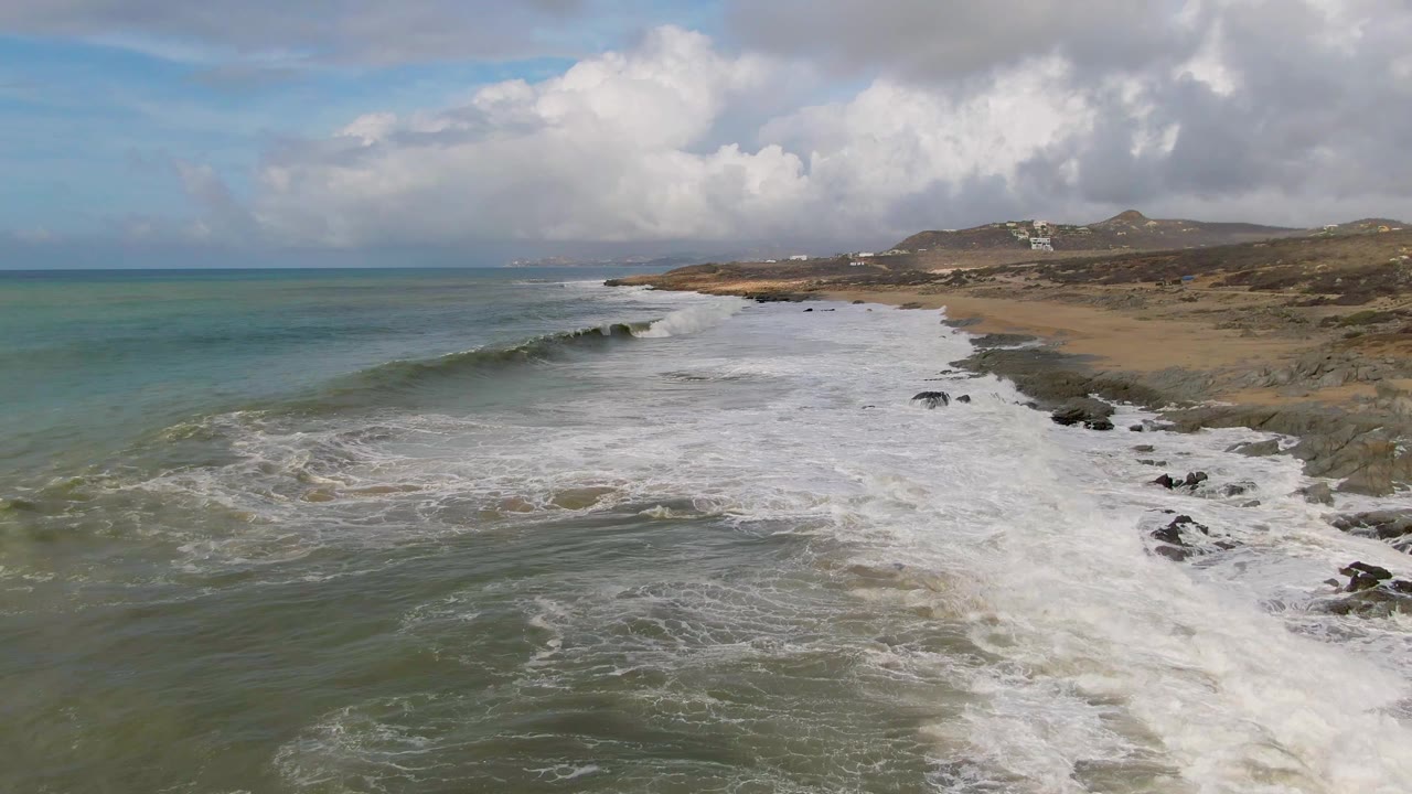 At 0:02, the Baja California Sur shoreline is covered by a wide sheet of white foam, with darker rocks breaking through near the surf edge and no visible people.