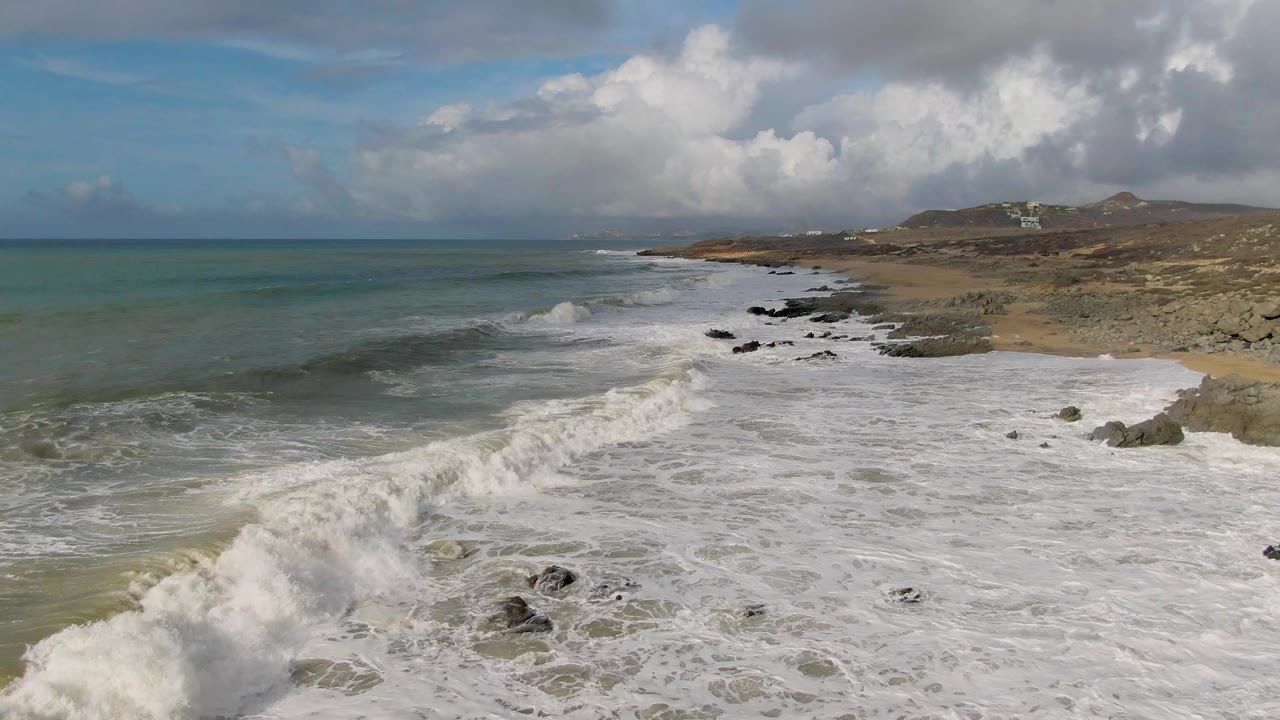 Aerial frame of Punta Gorda East Cape surf breaking across a sandy beach and scattered rocks, with green-blue water on the left, hills and distant buildings on the right, and no visible people.