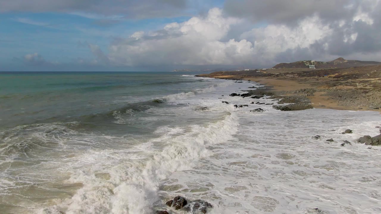 Aerial view over Punta Gorda East Cape as a larger band of white foam advances toward the foreground rocks, with open ocean to the left and no visible people on the beach.