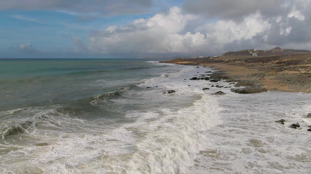 Punta Gorda East Cape coastline with whitewater spread wider across the beach, dark rocks breaking through the foam, distant hills under large clouds, and no visible people.