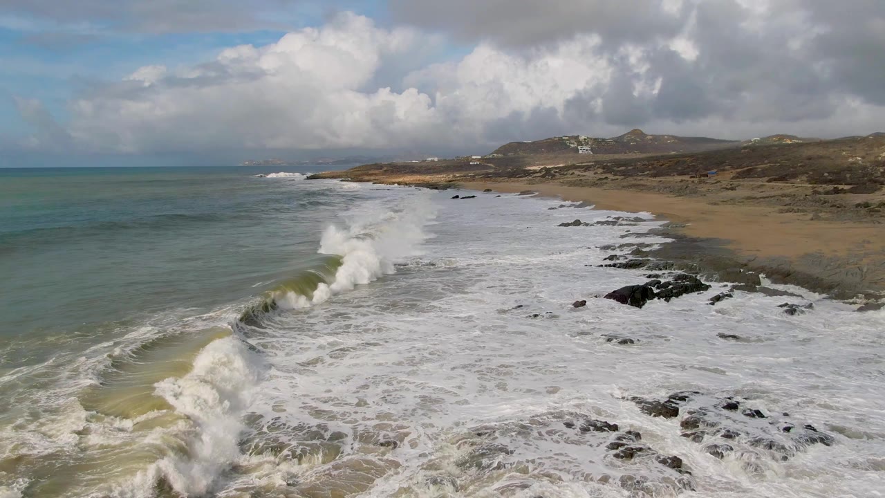 Aerial view at Punta Gordo, Baja California Sur, with a foamy wave breaking near dark rocky outcrops, sandy shore to the right, green-blue ocean to the left, and no visible people.