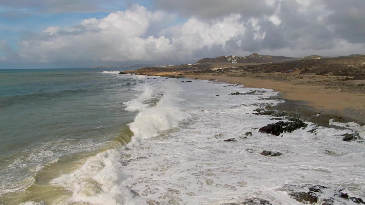 Large wave curls toward a sandy shoreline beside scattered rocks.