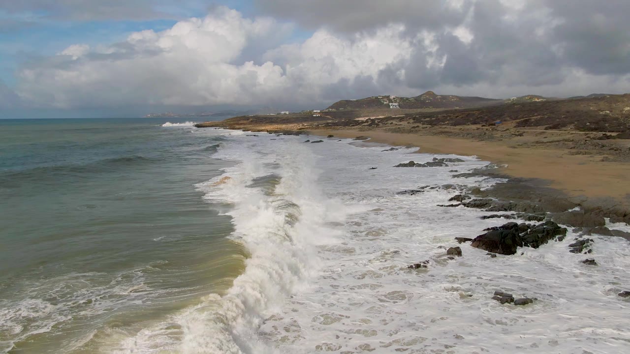 Foamy surf pushes farther down the Punta Gordo beach, with the green ocean on the left, rocks clustered on the right, low hills beyond the sand, and no visible people.