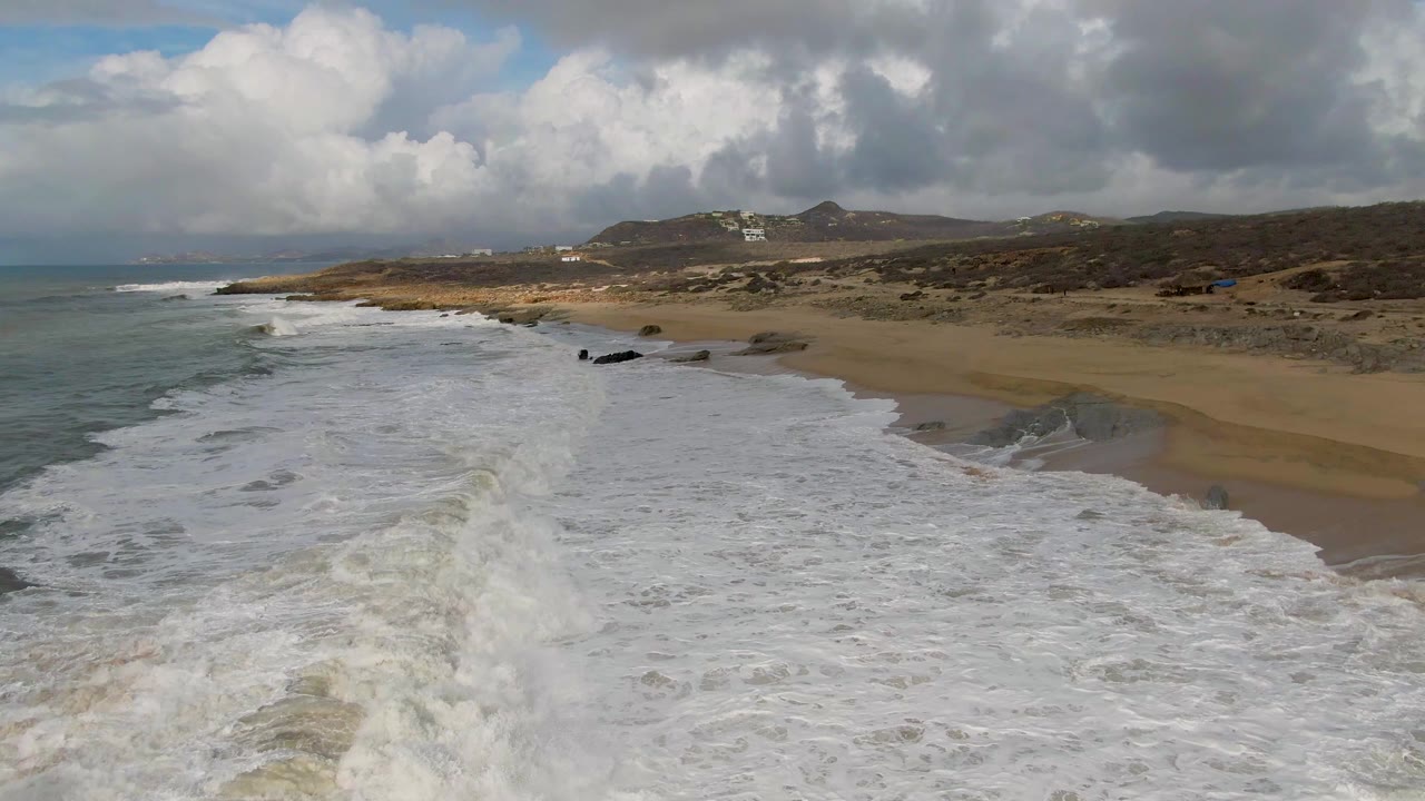 Aerial view of foamy waves reaching a sandy beach with rocks and hills beyond.