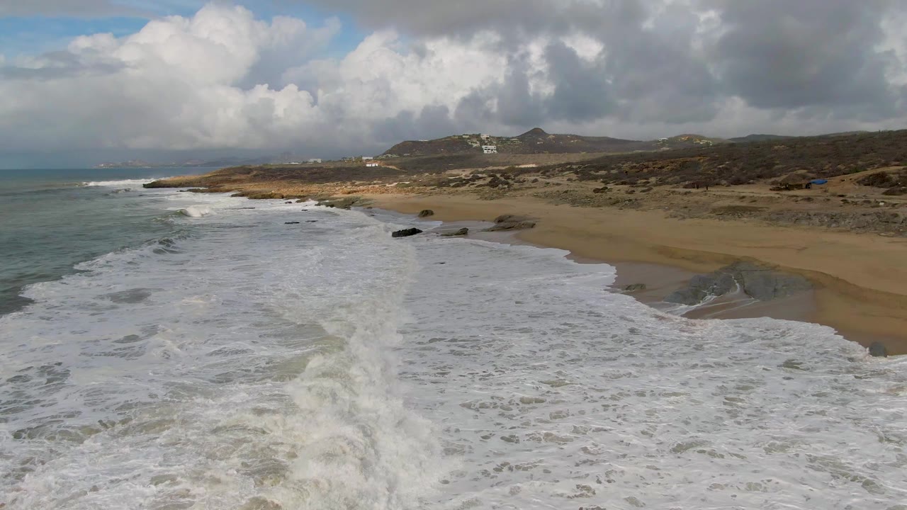 Aerial view at Punta Gordo with a diagonal band of breaking waves, tan beach on the right, rocky patches near the waterline, distant hills, and no visible people.