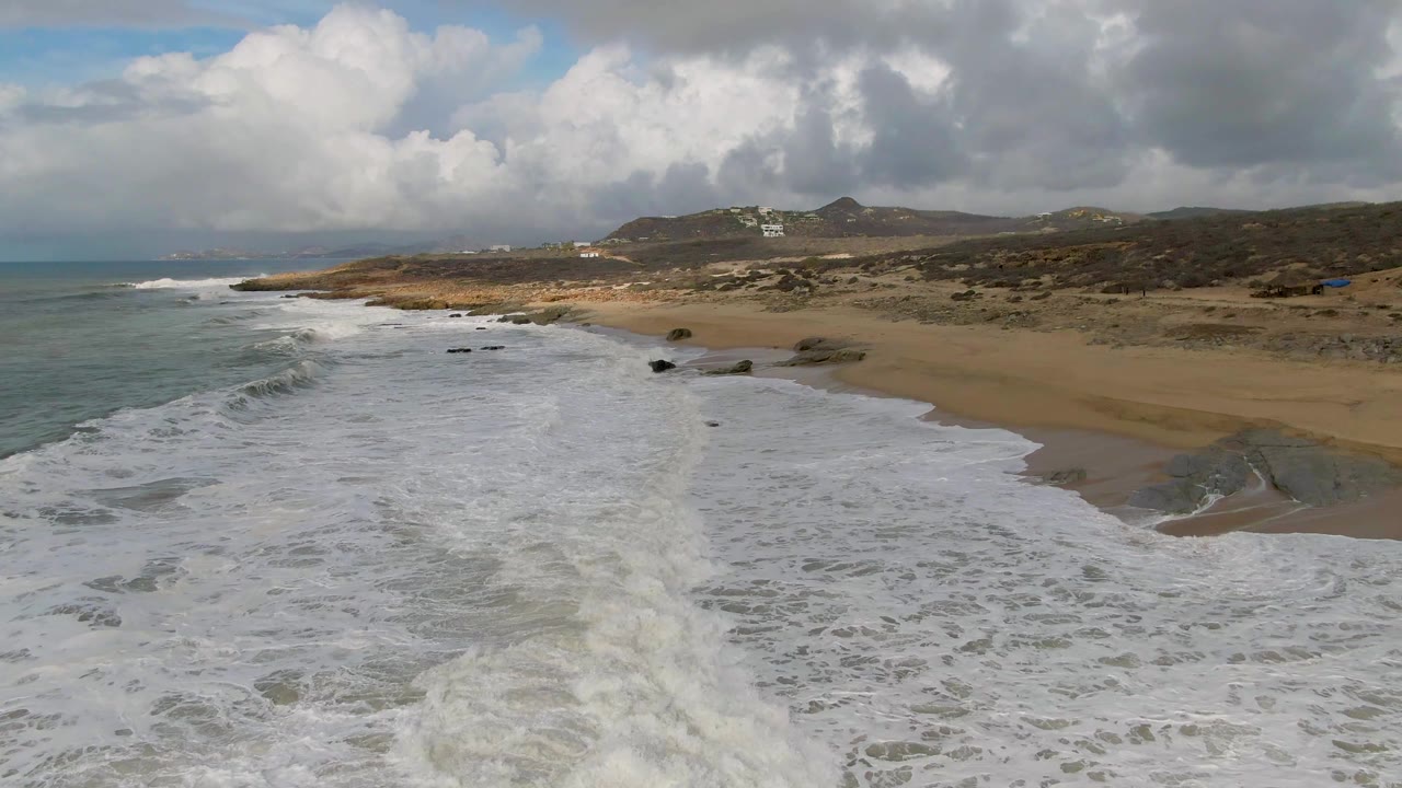 Punta Gordo shoreline frame with foamy water filling the lower half, scattered dark rocks near the beach, brown hills in the distance, layered clouds overhead, and no visible people.