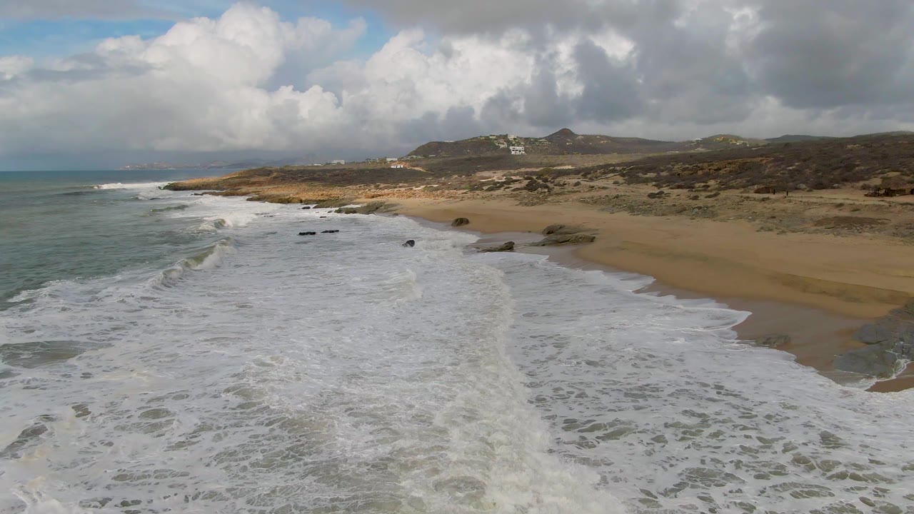 Aerial coastal frame from Punta Gordo showing a fresh wave line on the left, foam across the foreground, a curving sandy beach to the right, low hills beyond, and no visible people.