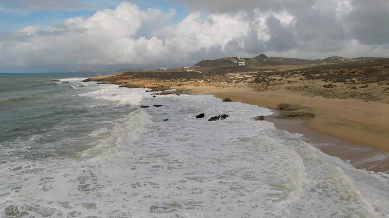 Aerial view at Punta Gorda East Cape, Baja California Sur, with broad white surf covering the foreground, dark rocks near the shoreline, dunes and low hills beyond, and no visible people.