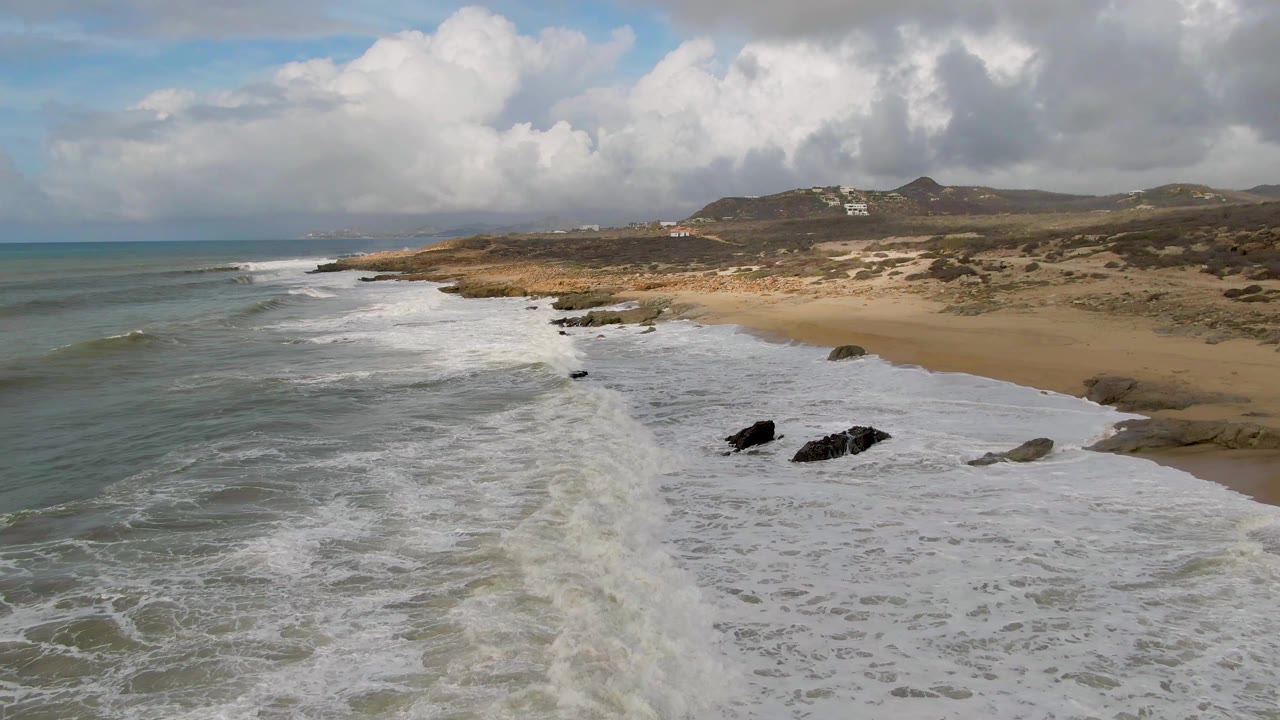 At 0:02, the whitewater line stretches diagonally toward the sandy Punta Gorda East Cape beach, with teal-gray ocean to the left, rocks in the wash, dunes behind, and no visible people.
