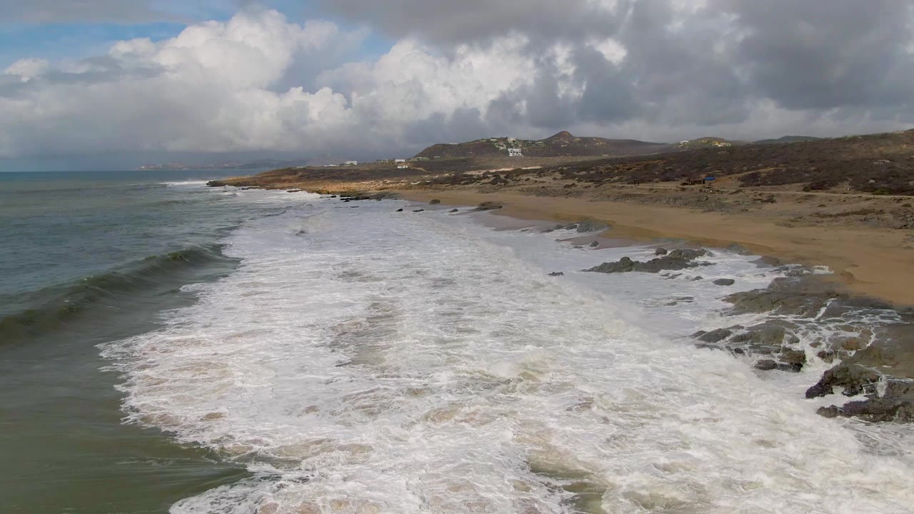 Aerial view of surf washing onto a sandy beach with rocks and cloudy sky