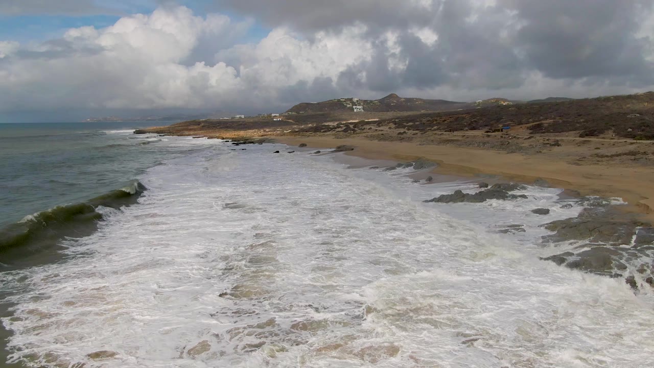 Aerial view at Punta Gorda with a darker wave face forming along the left edge, broad whitewater across the center, sandy beach and dark rocks to the right, distant hills, clouds, and no visible people.
