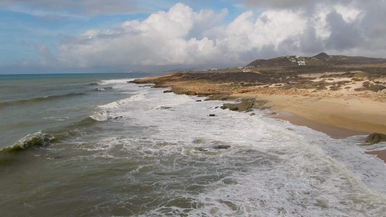 Aerial view at Punta Gorda, Baja California Sur, with green-blue waves rolling toward a sandy beach and dark rocks under a cloud-filled sky, no visible people.
