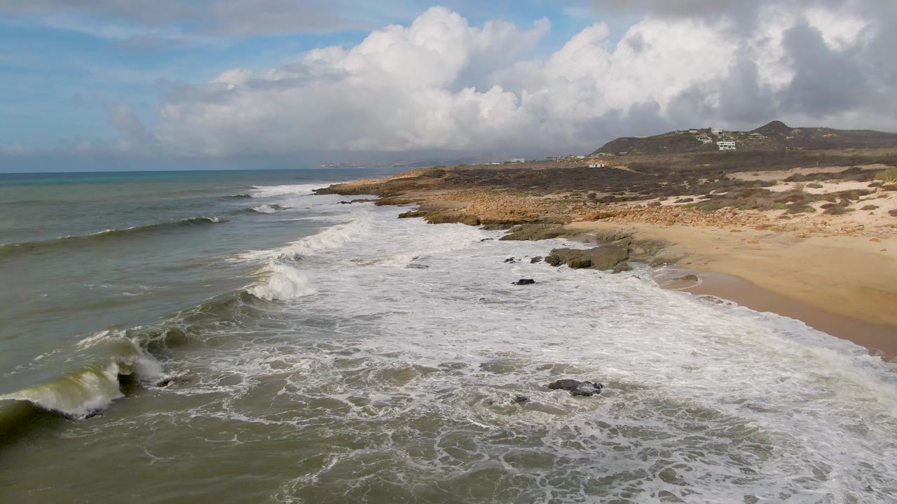 At Punta Gorda, a larger wave curls in the left foreground while white foam spreads over rocks and sand, with low hills and clouds in the distance and no visible people.