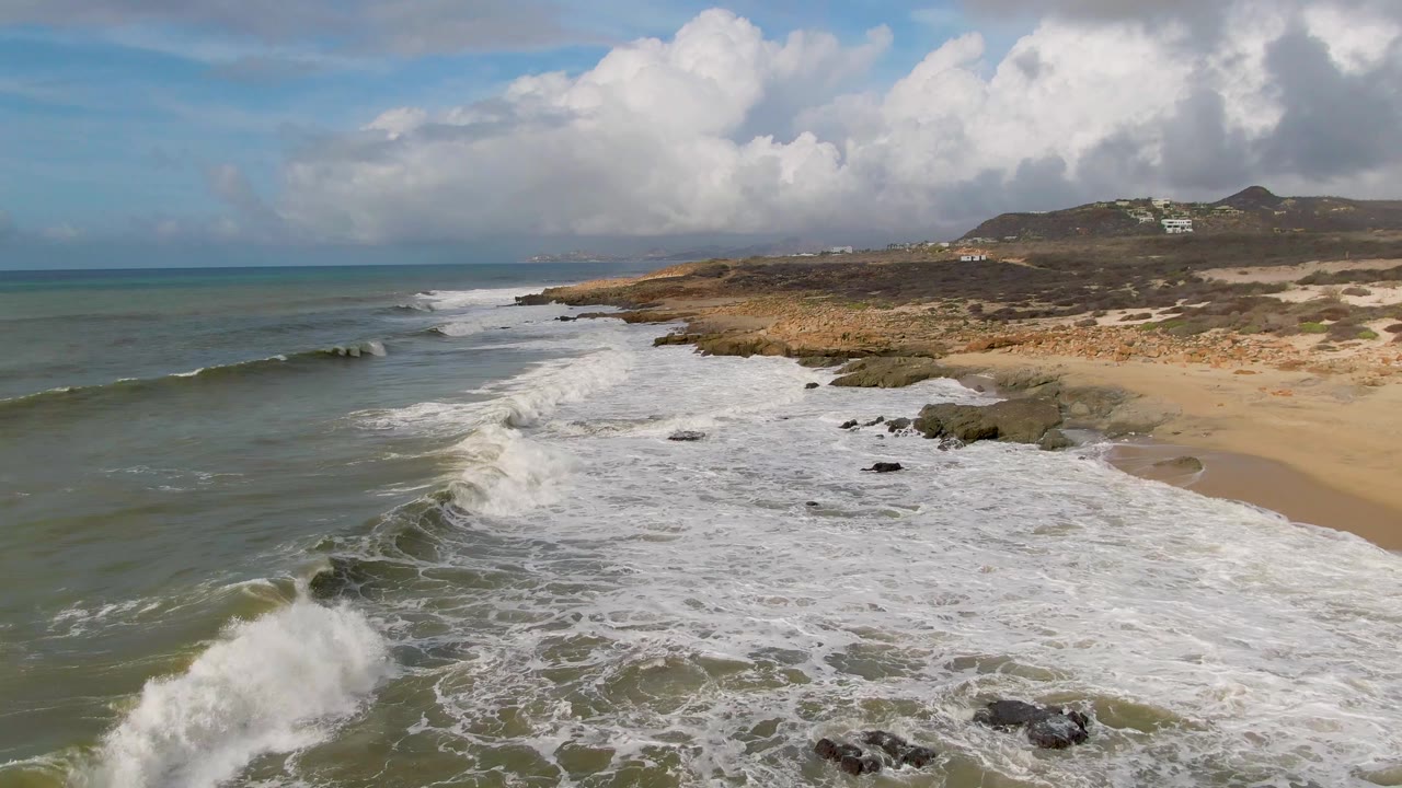 A wave breaks near the lower foreground at Punta Gorda as foam runs across scattered rocks and pale sand, with open water to the left and no visible people.