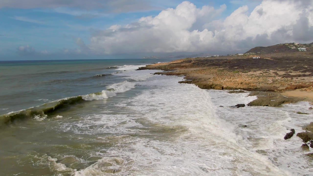 Aerial view at Punta Gorda East Cape, Baja California Sur, with white waves breaking diagonally toward a rocky tan shoreline, green-blue ocean at left, distant hills, and no visible people.