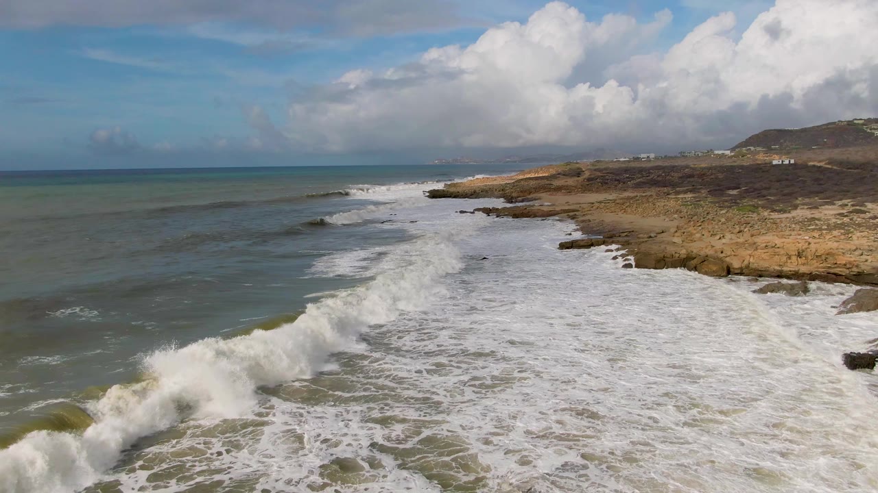 White surf breaks harder across the foreground rocks at Punta Gorda East Cape, with a broad foam field, green ocean beyond, tan shoreline at right, distant hillside, and no visible people.