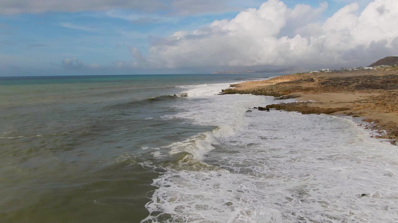 Aerial coastal frame from Punta Gordo showing a breaking wave in the midground, white foam covering the right side of the shallows, tan rocky coast beyond, distant clouds, and no visible people.