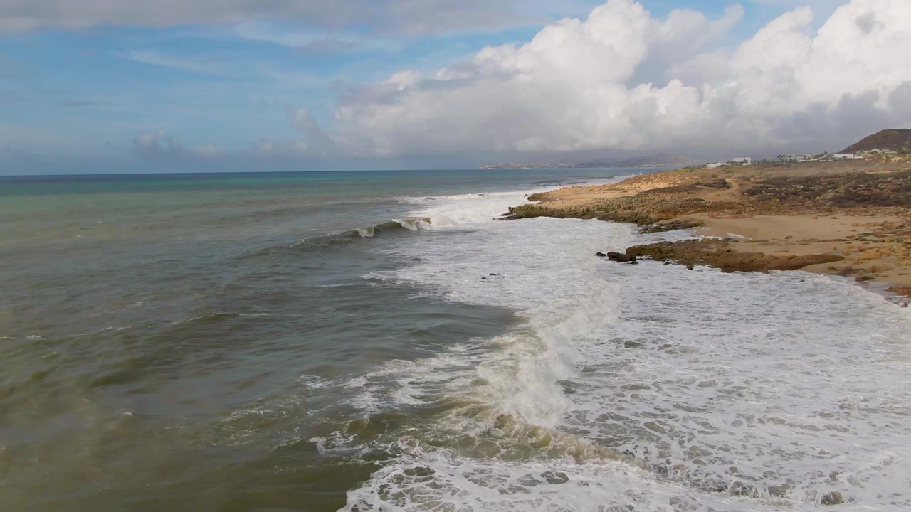 Punta Gordo shoreline frame with whitewater spreading across shallow green water, darker ocean to the left, scattered rocks near the surf, tan shore to the right, bright clouds overhead, and no visible people.