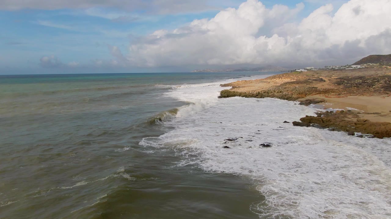 Aerial frame of surf spreading across rocks and shallow water near Punta Gorda, Baja California Sur, with tan coast, distant hills, and no visible people.