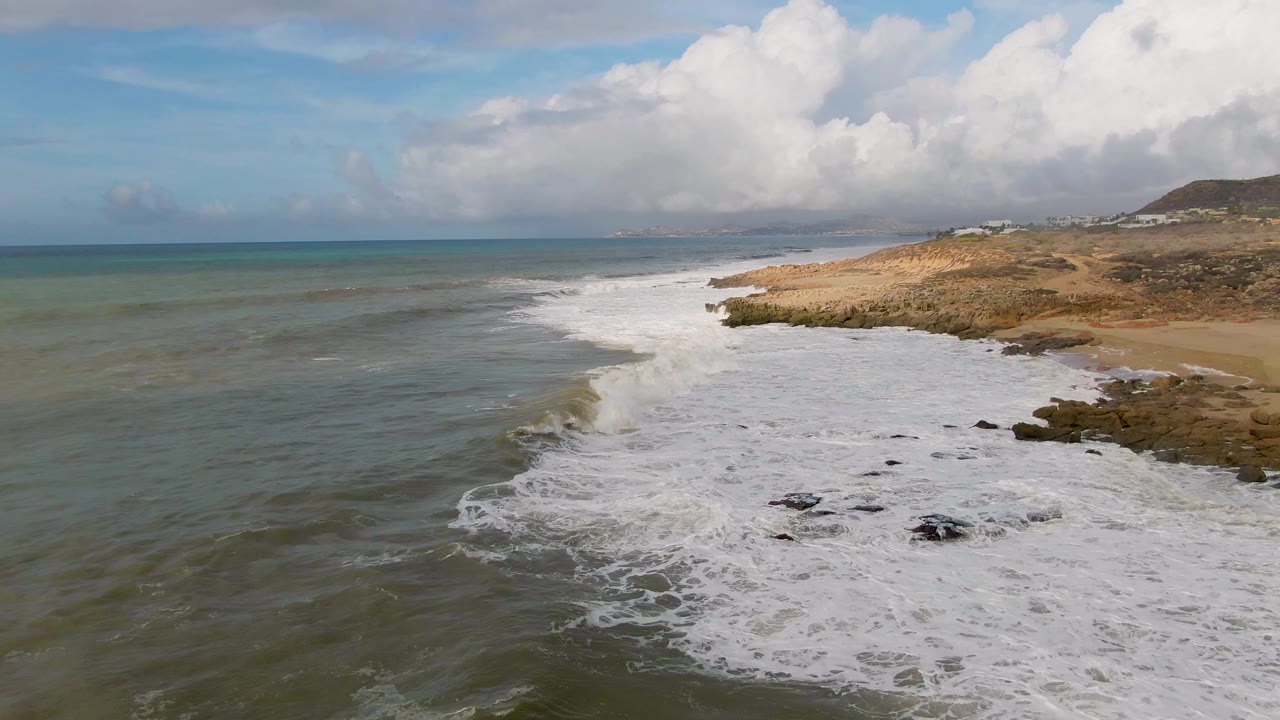 Surf advances across dark rocks in green-blue water near Punta Gorda, with the coastline running along the right side and no visible people.