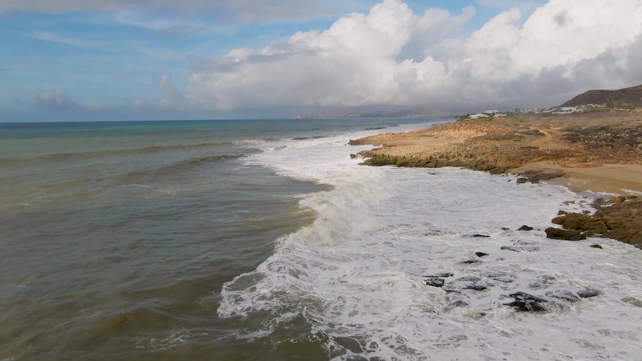 A wider patch of white foam covers the nearshore rocks at Punta Gorda while shallow green water fills the left side and no visible people appear on the coast.