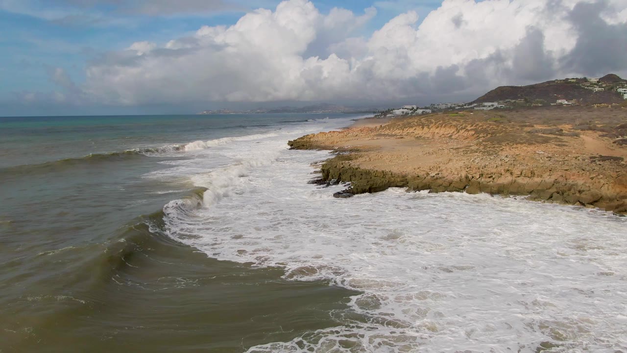 A darker wave face lifts in the foreground ocean at Punta Gorda while older foam spreads against the rocks and the tan shoreline continues beneath a cloud-filled sky, with no visible people.