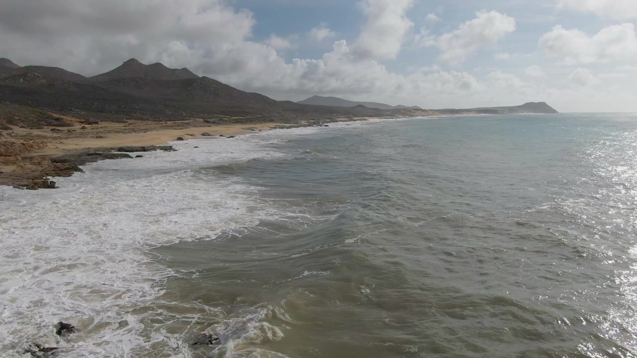 Aerial view of surf washing onto a sandy shoreline below dark mountains.
