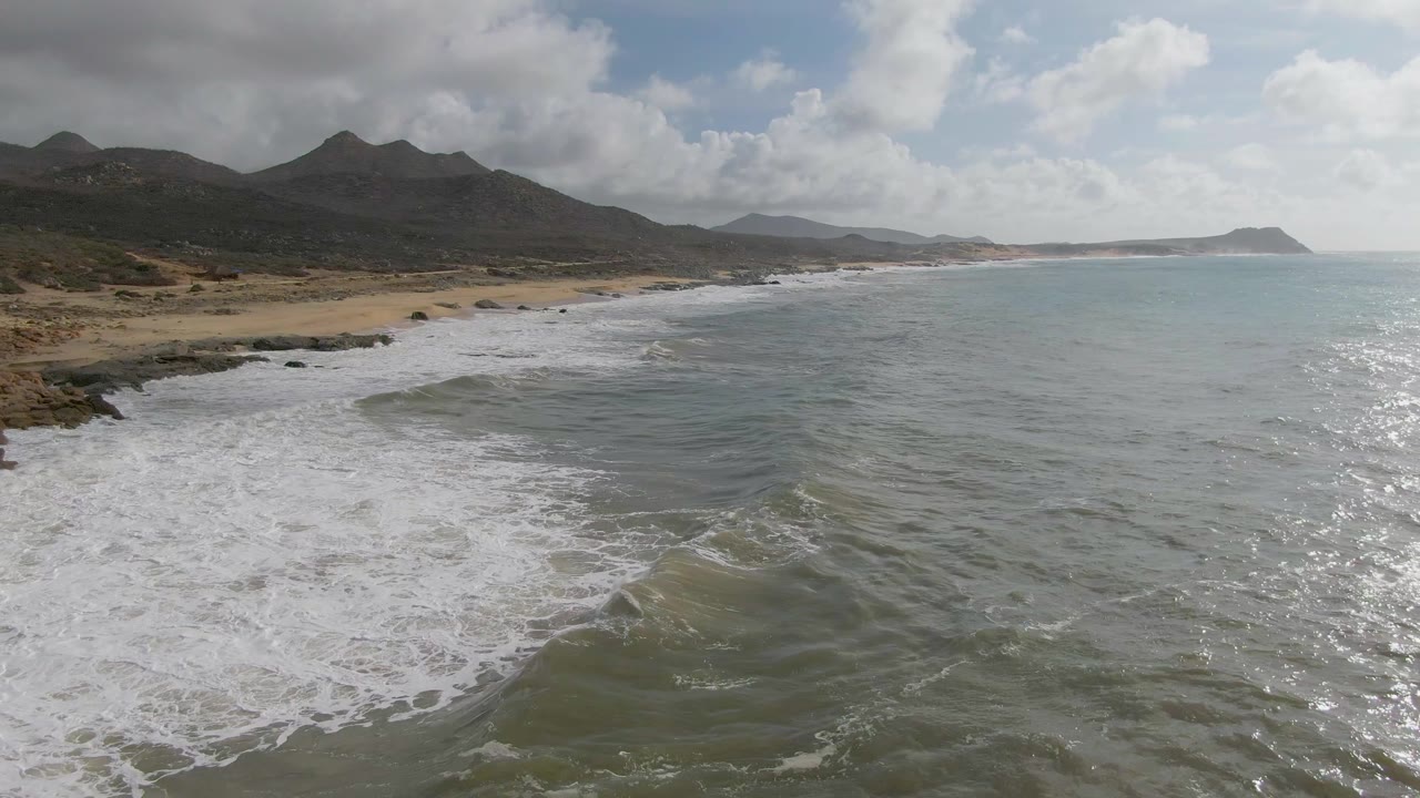 Punta Gorda East Cape coast seen from above with breaking white waves along the left shoreline, calmer green-blue water to the right, mountain ridges behind, and no visible people.