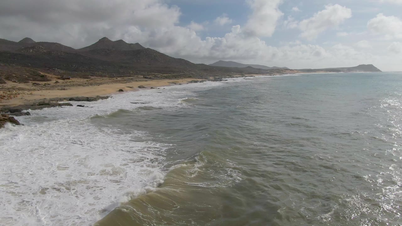 Punta Gorda East Cape beach with receding white foam in the lower left, open water across the foreground, mountains along the horizon, and no visible people on the sand or in the water.