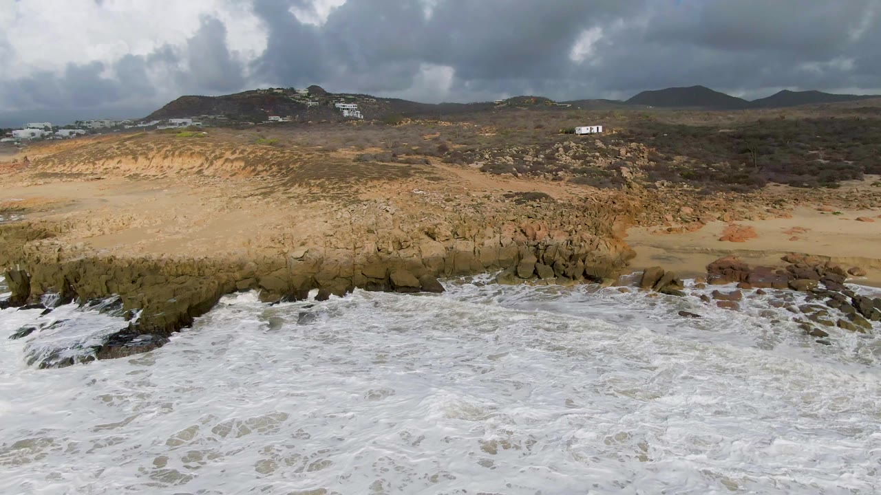 Aerial frame over Punta Gorda, Baja California Sur, showing white surf in the foreground, a rocky sandy shoreline, low hills, heavy clouds, and no visible people.