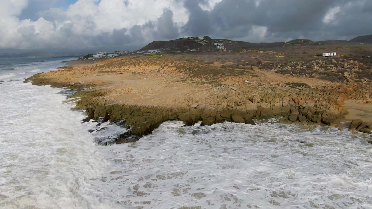 Aerial view in Punta Gorda, Baja California Sur, with foamy water curling around dark coastal rocks, sand behind the break, hills under cloud, and no visible people.