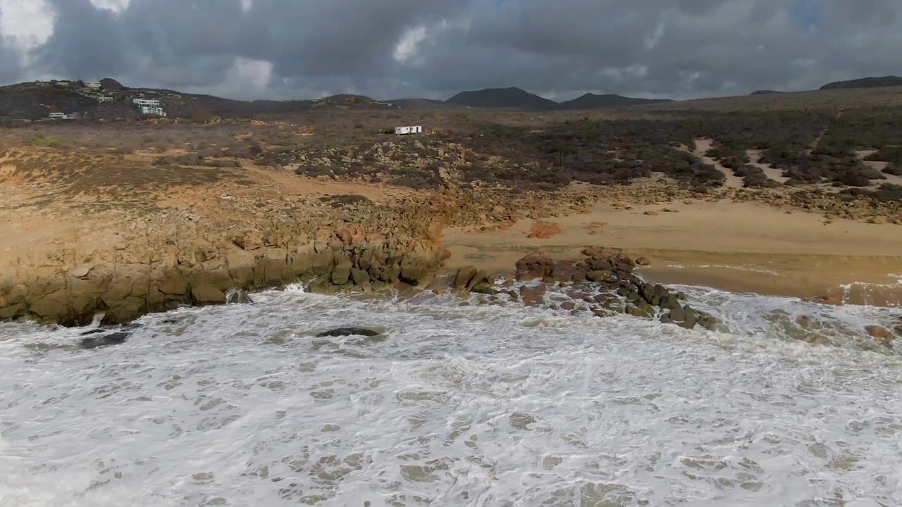 Rocky beach with white surf in front and darker hills under clouds