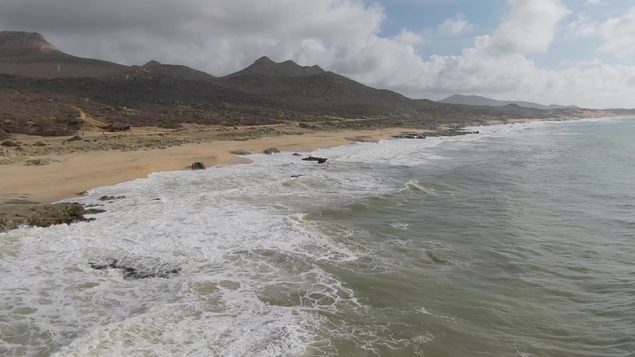 Aerial view of waves breaking on a sandy rocky shoreline below mountains.