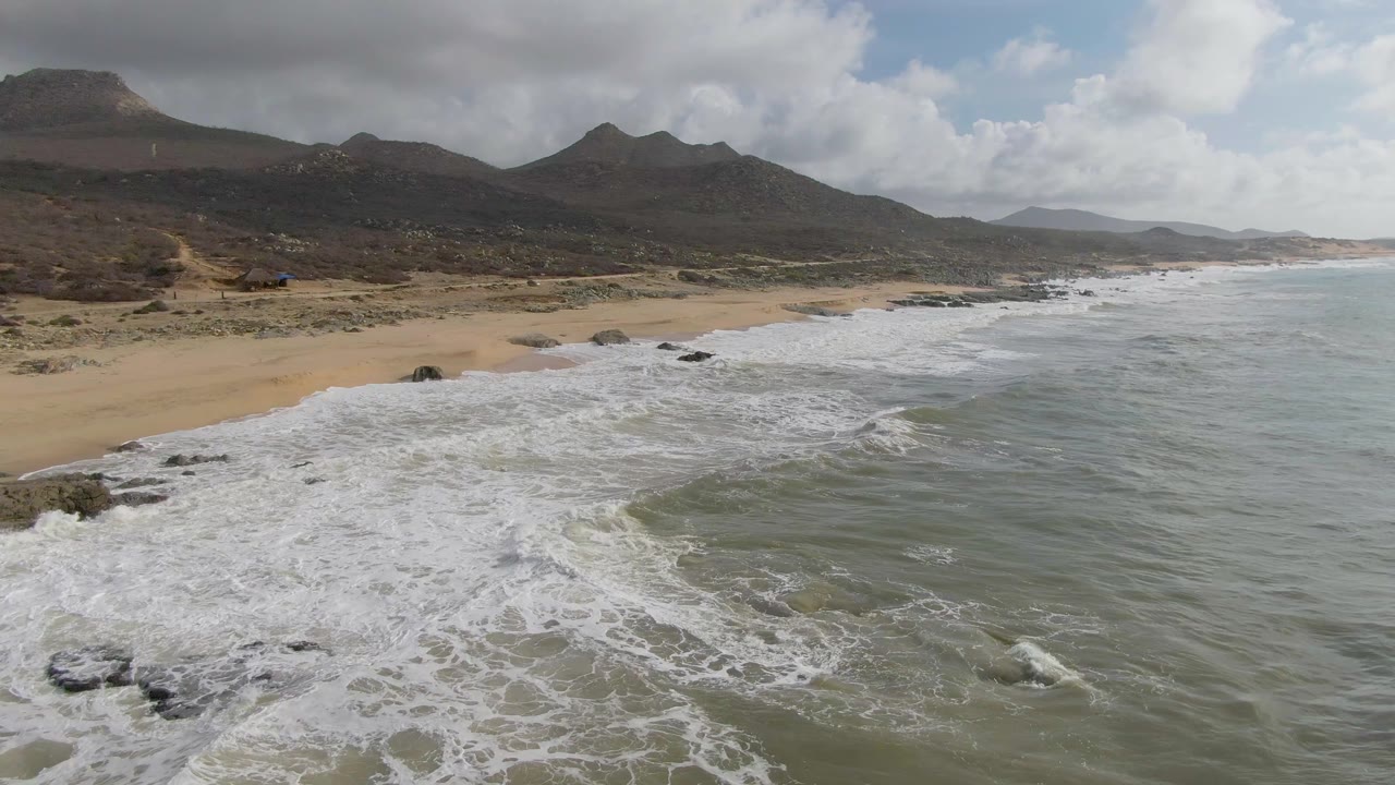 At 0:01, white water pushes farther along the rocky Punta Gorda shore while the sandy beach curves below dark mountains, with no visible people.