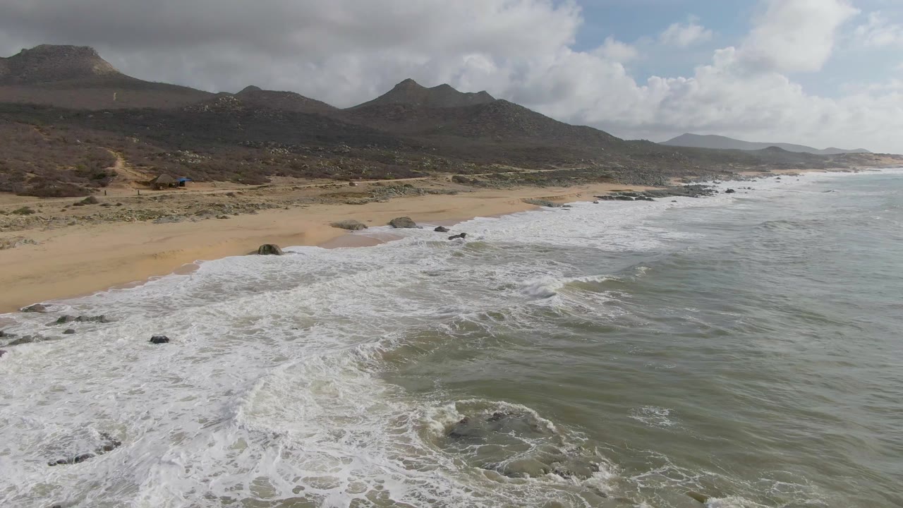 At 0:02, churning surf wraps around scattered rocks off Punta Gorda while a long sandy beach and layered mountains remain visible, with no visible people.